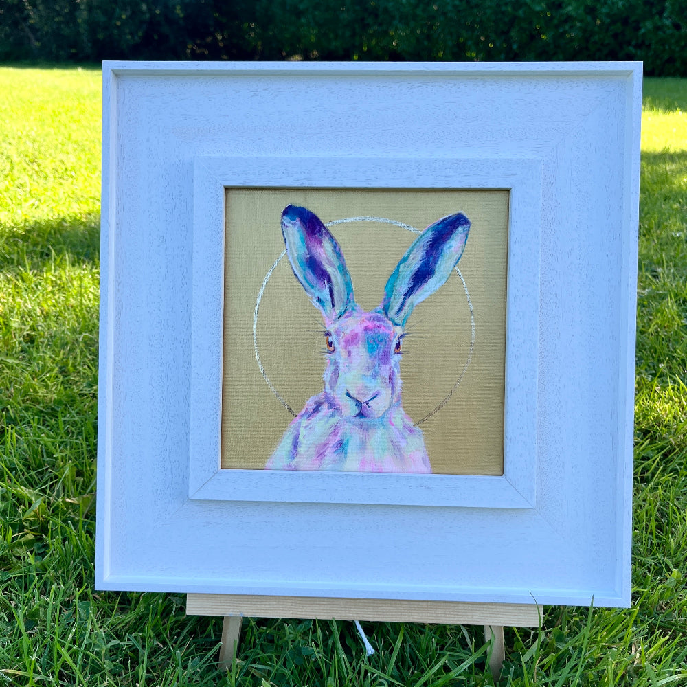 A framed art print of a colorful painted hare on a tan background, displayed on a small easel outdoors on a green grass lawn with trees in the background.
